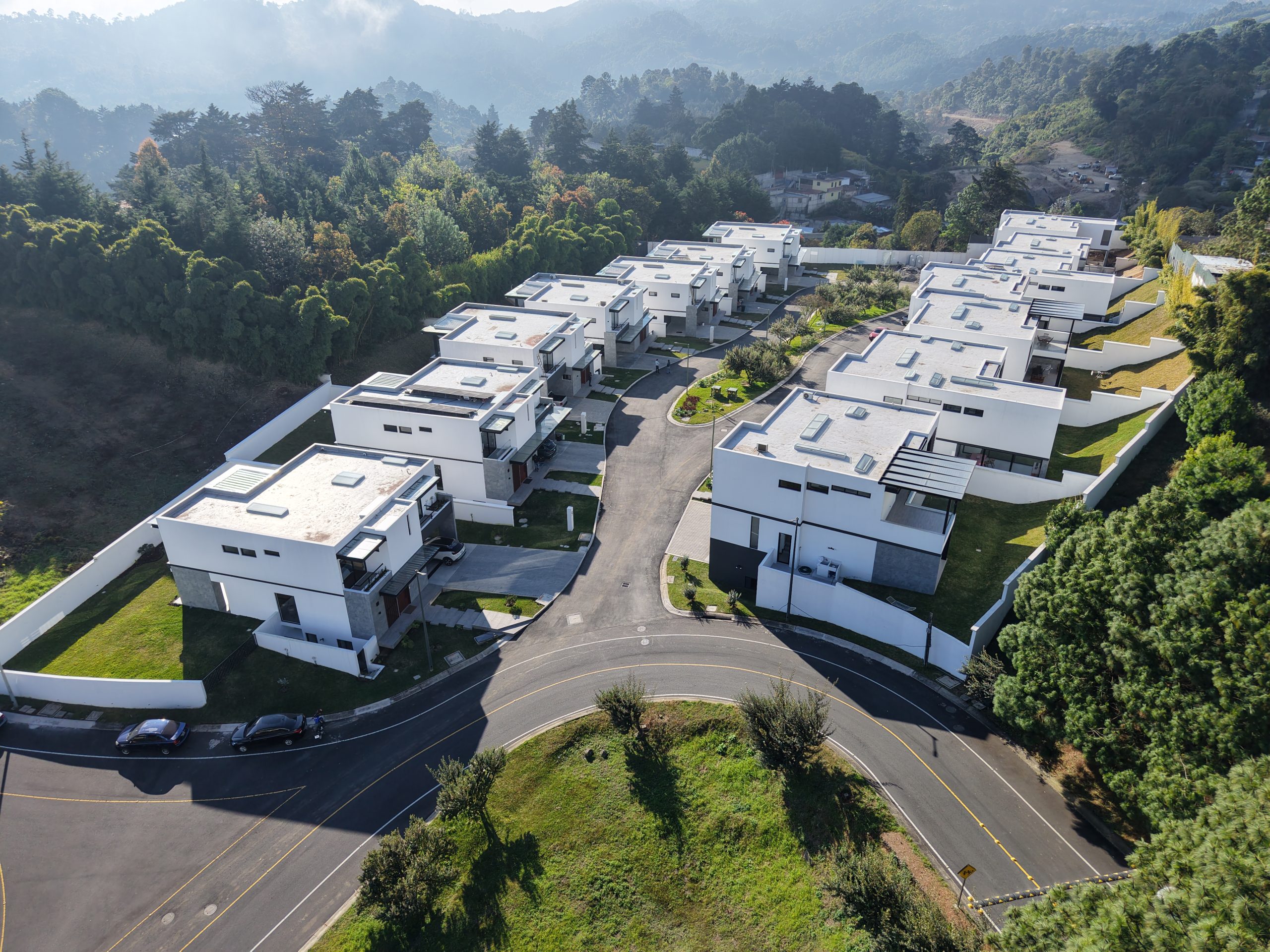 Aerial view of a modern white townhouse complex arranged along a curved road amid green lawns and trees