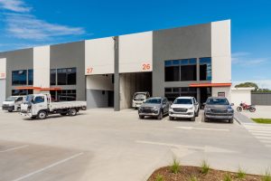 Front view of a modern industrial building with bays 26 and 27 gray and white panels and parked trucks and cars in the lot