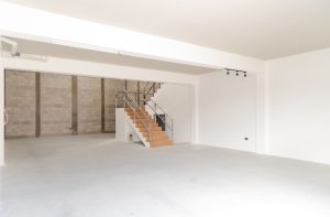 Interior of an empty warehouse or showroom with white walls concrete floor and a wooden stairway with metal railing leading to an upper level