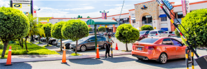 Parking lot in front of a shopping center with trimmed trees orange traffic cones and a barrier arm a worker stands near the barrier beside an orange car under a sunny sky