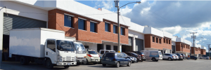 Row of brick industrial warehouses with loading bays and a line of parked trucks and cars outside the buildings