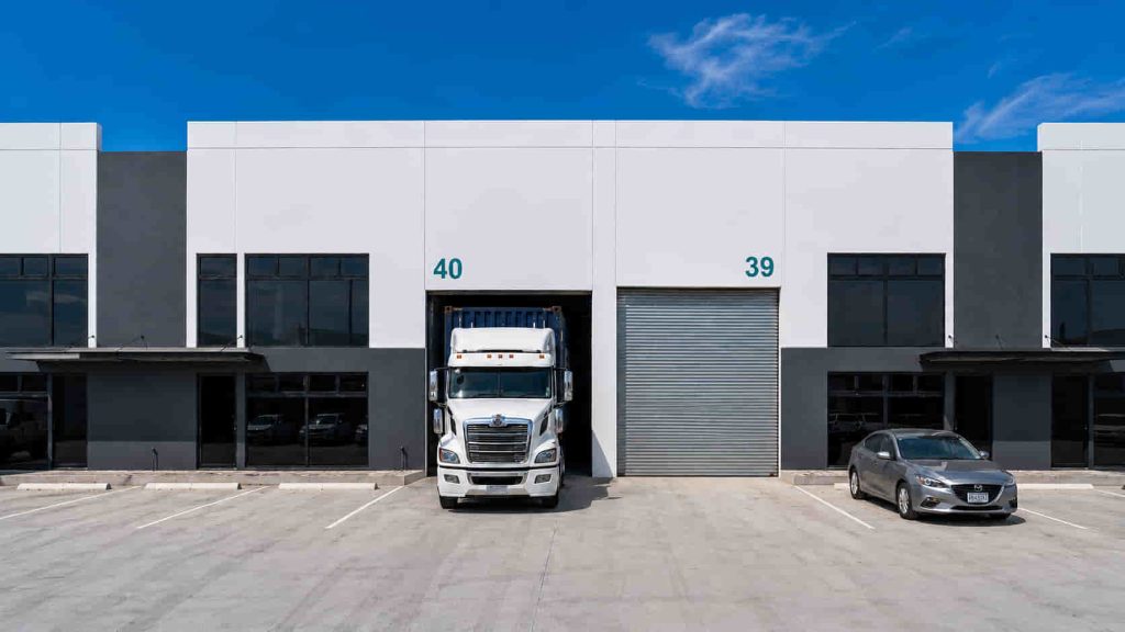 White semi-truck entering loading bay 40 of a modern warehouse, with a gray car parked to the right and a blue sky above.