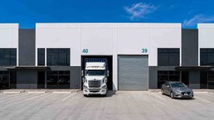 White semi truck entering loading bay 40 of a modern warehouse with a gray car parked to the right and a blue sky above