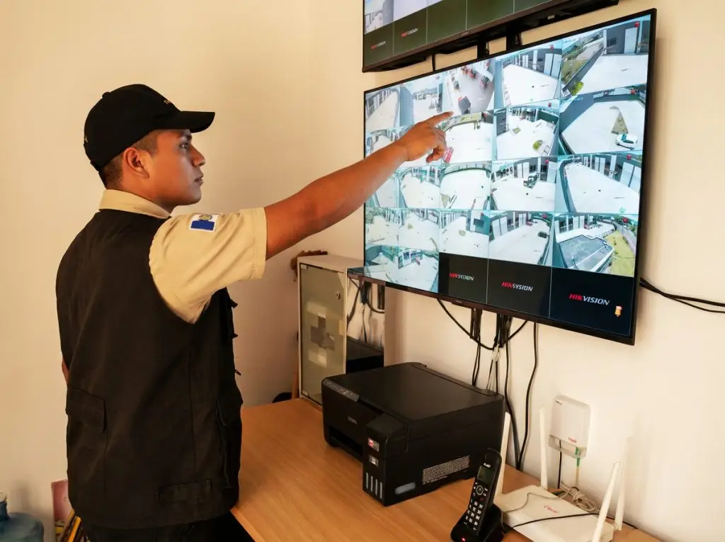 Security guard points at a wall of cctv monitors displaying multiple camera feeds